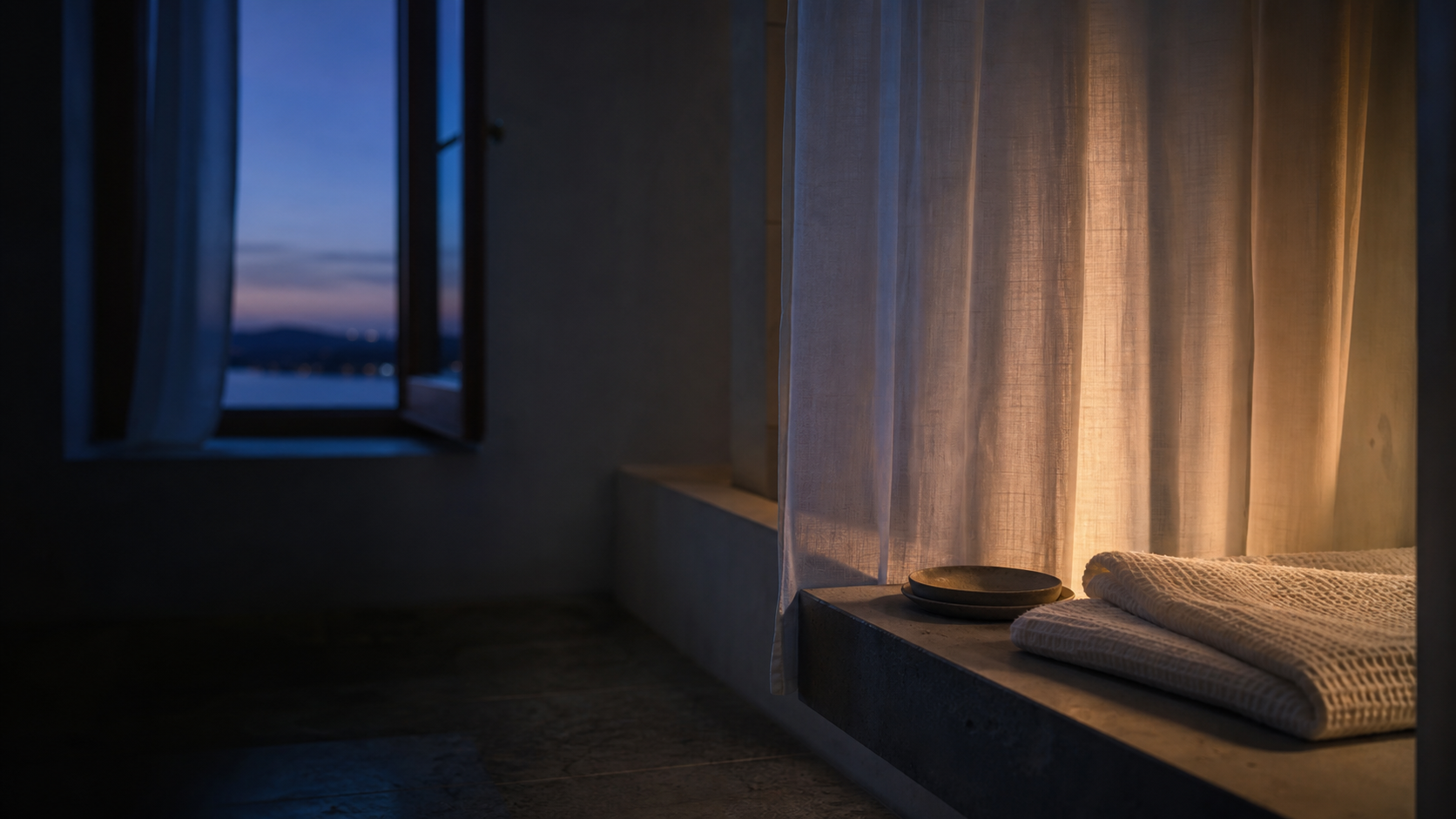 A quiet bathroom at blue hour with folded white textiles and warm light